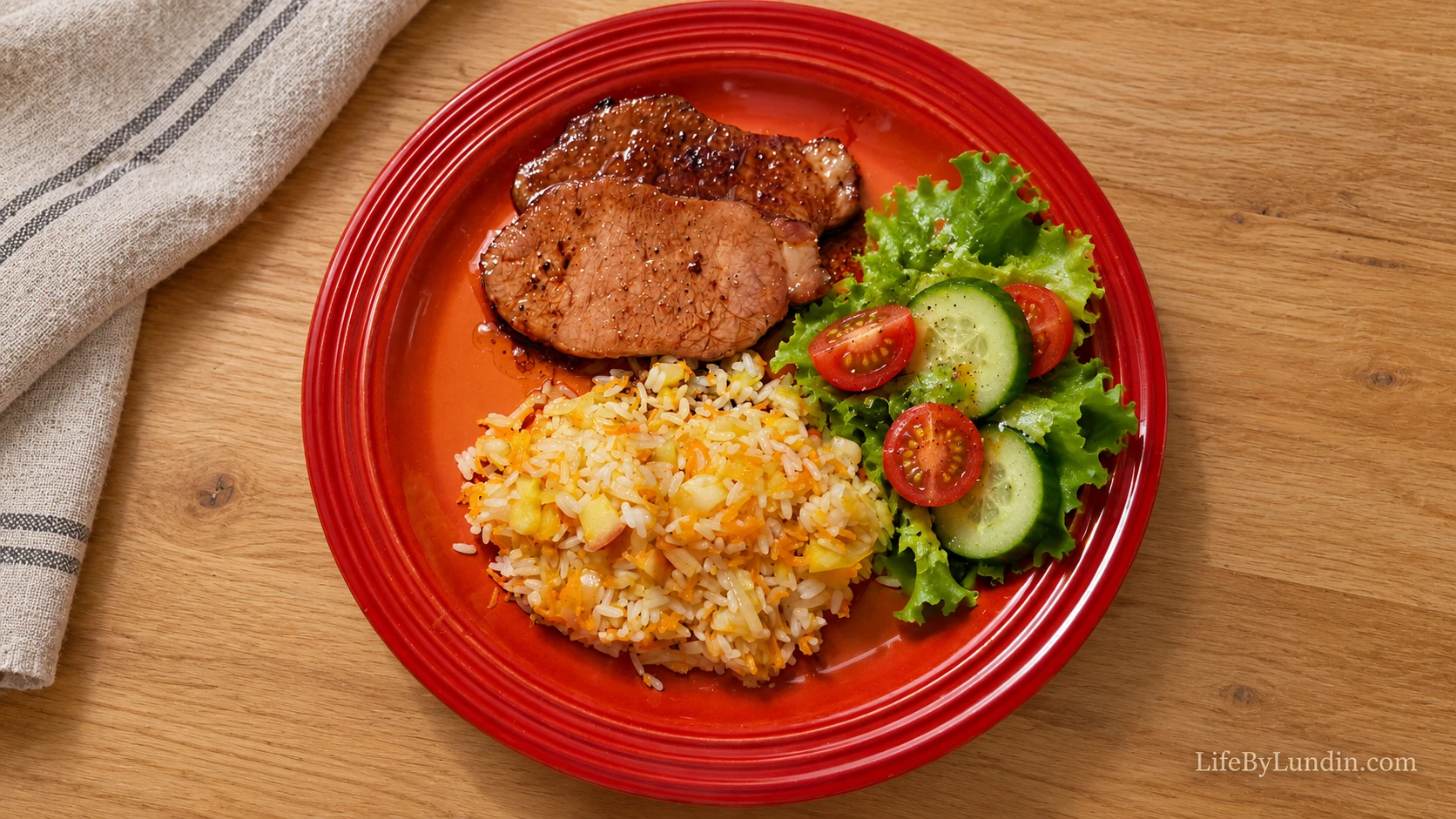 Honey-glazed smoked pork and apple and onion rice with lettuce, cucumber and tomato on a red plate on a light wooden table