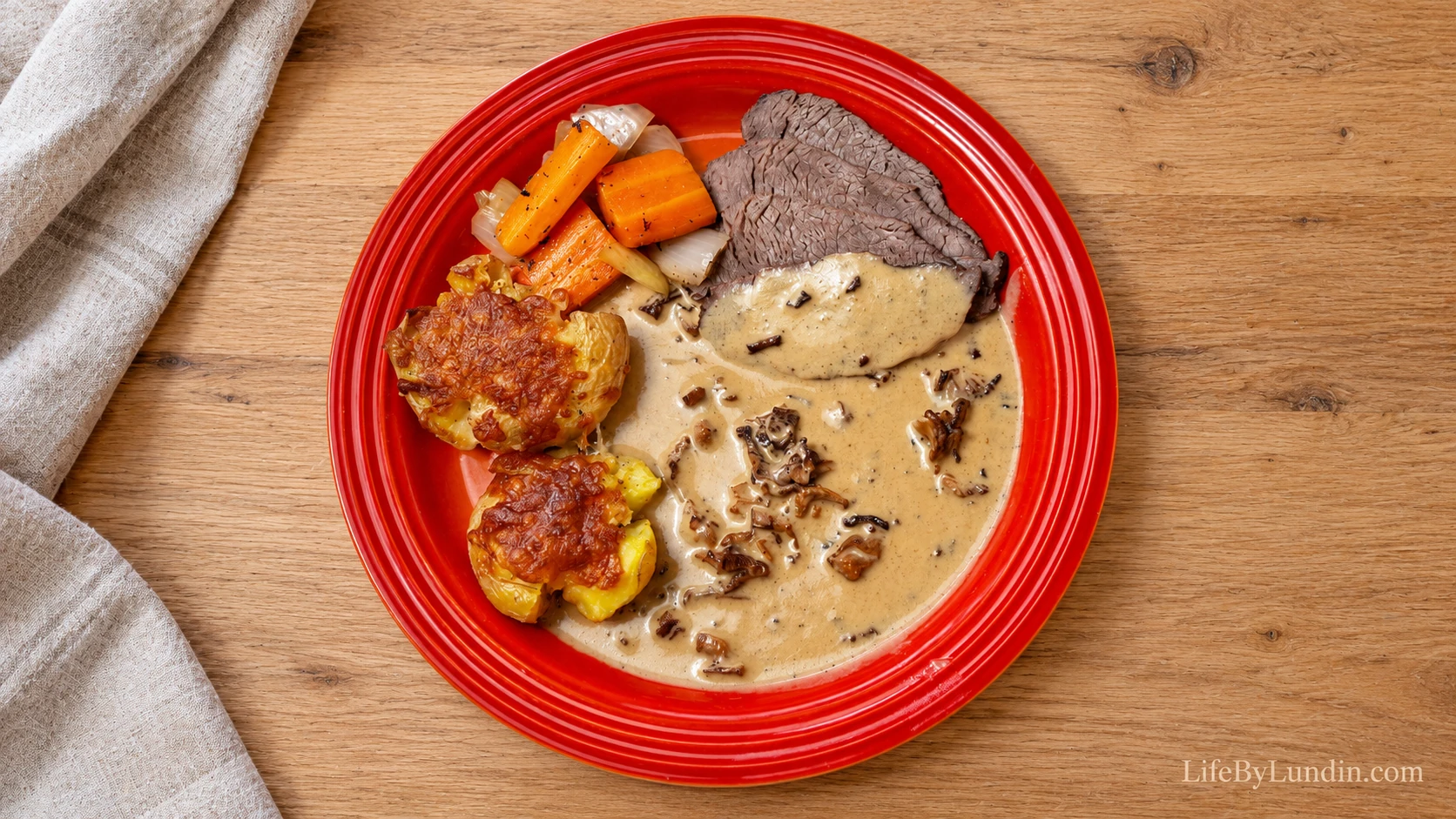 Sliced roast beef, cheesy smashed potatoes, carrot and onion with chanterelle cream sauce on a red plate on a light wooden table