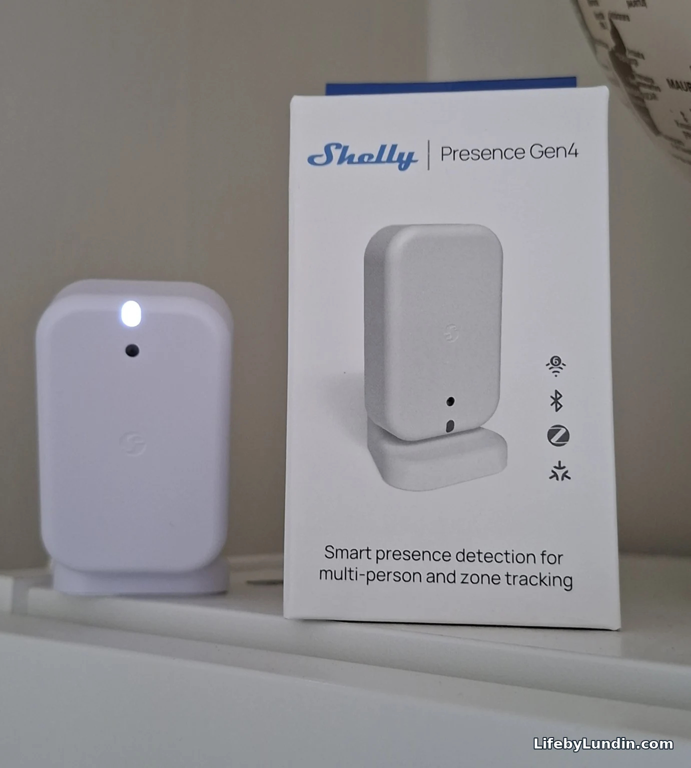 Vit Shelly Presence Gen4 på sitt stativ bredvid produktförpackningen